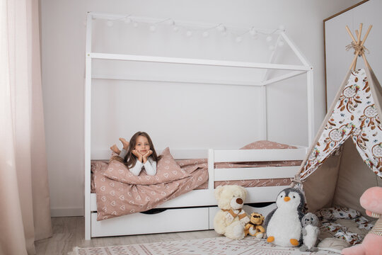 Portrait of a beautiful caucasian little girl smiling while relaxing in her bedroom during a leisure day. Cheerful female kid lying on scandinavian wooden house frame bed.