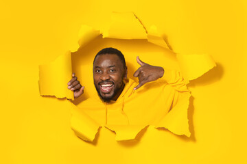 Handsome african american man making phone gesture, posing through breakthrough of yellow paper background
