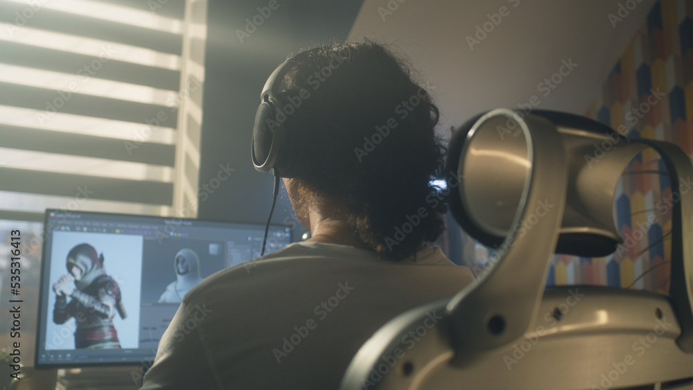 Poster Back view of man in headphones sitting at the table and creating video game hero and 3D animation in professional computer program, while working remotely at home.