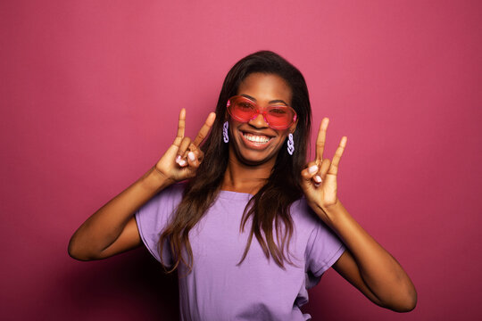 Joyful excited rock fan shouting and showing devils horn hand gesture. Beautiful African American lady standing isolated over pink background.