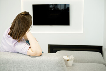 A woman is sitting on a sofa, watching TV, Asian food in a box is nearby, a lonely evening.