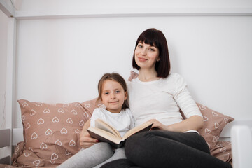 Close up of smiling mom and little daughter relax in cozy bed read funny interesting children book together, happy mother and small preschooler girl child enjoy fairytale rest in comfortable bedroom