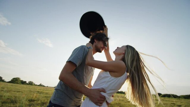 Loving couple kissing and cuddling against summer sunset. Loving couple kisses and cuddles among green field. Lady with long straight hair embraces bearded boyfriend in sunglasses against summer