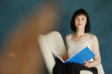 Young self confident woman with short hair sitting, relaxing on chair and holding blue folder and checking documents. Successful brunette female. Education, business, lifestyle concept