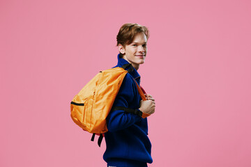 a happy guy in a blue sweater stands sideways to the camera with a orange backpack on his back....