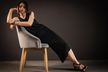Fototapeta na wymiar Studio portrait of a tired, tortured, young beautiful sensual woman in a black dress, sitting on a wooden chair, leaning on the back, on a dark background. Depression