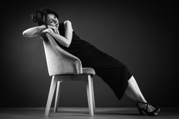 Obraz na płótnie Canvas Studio portrait of a tired, tortured, young beautiful sensual woman in a black dress, sitting on a wooden chair, leaning on the back, on a dark background. Depression