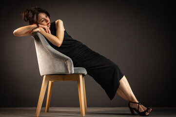 Studio portrait of a tired, tortured, young beautiful sensual woman in a black dress, sitting on a wooden chair, leaning on the back, on a dark background. Depression