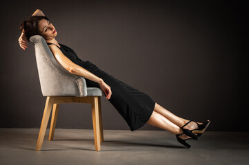 Fototapeta na wymiar Studio portrait of a tired, tortured, young beautiful sensual woman in a black dress, sitting on a wooden chair, leaning on the back, on a dark background. Depression