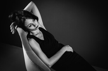 Studio portrait of a tired, tortured, young beautiful sensual woman in a black dress, sitting on a wooden chair, leaning on the back, on a dark background. Depression