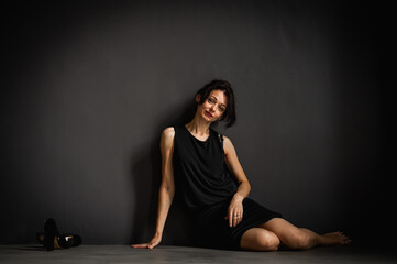 Fototapeta na wymiar Studio portrait of a tired, tortured, young beautiful sensual woman in a black dress sitting on the floor on a gray background. Depression, fatigue, loneliness, thoughtfulness