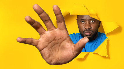 Serious african american man reaching through hole in torn yellow paper, trying to grab something - obrazy, fototapety, plakaty
