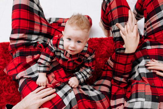 Happy young parents and baby boy in the same pyjamas lying on red floor carpet and smiling at camera, top view. family relax at home, enjoying time together on holiday. Xmas and New Year. Valentines