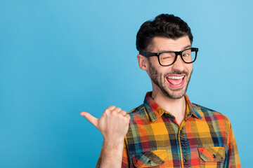 Photo of friendly handsome positive guy in eyewear plaid shirt indicating at empty space blinking isolated on blue color background