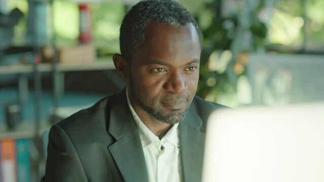 Handsome african american man working on the computer looking on screen serious in the office at his desk. Sunlight. Corporate. CEO. Businessman. Slow motion