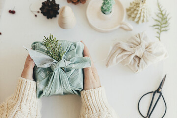 Zero waste Christmas concept. Hands holding gift box wrapped in green fabric with fir branch on white wooden table with eco friendly decor and candle. Furoshiki gift wrapping. Eco winter holiday