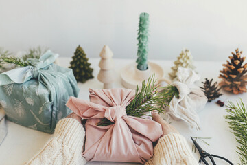 Furoshiki gift wrapping. Hands holding gift box wrapped in pink fabric with fir branch on white wooden table with eco friendly decor and candle. Zero waste Christmas concept.  Merry Christmas!