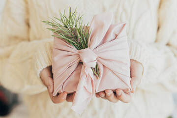 Zero waste Christmas. Hands holding gift wrapped in pink fabric with fir branch. Stylish Furoshiki...