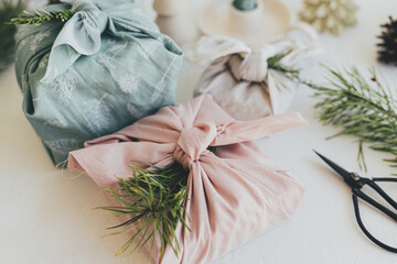 Zero waste Christmas concept. Stylish wrapped gifts in linen fabric on white rustic table with eco friendly wooden toys, fir branches, candle. Furoshiki gift wrapping. Happy Holidays!