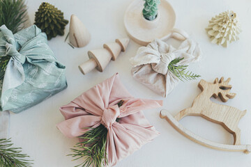 Zero waste Christmas flat lay. Stylish wrapped gifts in linen fabric on white rustic table with eco wooden tree, deer, fir branches, candle. Furoshiki gift wrapping. Eco friendly toys.