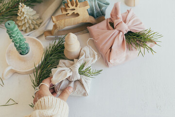 Zero waste Christmas. Hand holding gift box wrapped in linen fabric with fir branch on background of white table with eco friendly wooden decor and candle. Furoshiki gift wrapping. Merry Christmas!