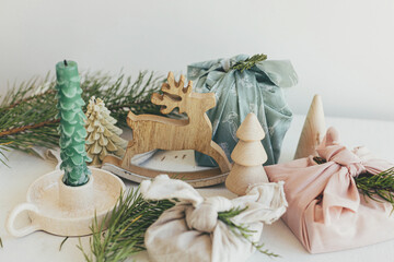 Merry Christmas!  Stylish wrapped gifts in linen fabric on white rustic table with eco wooden tree, deer, fir branches, candle. Furoshiki gift wrapping. Eco friendly toys. Zero waste Christmas