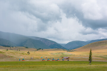 Scenic view from sunlit grassy steppe to small houses on background of green high mountain range in rainy clouds. Dramatic landscape with meadow and small lodges in mountains at changeable weather.