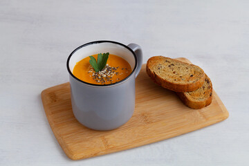Vegetarian autumn soup. Pumpkin cream soup in a grey cup on a cutting board. copy space. Mug of tasty pumpkin soup with crackers on the white background.