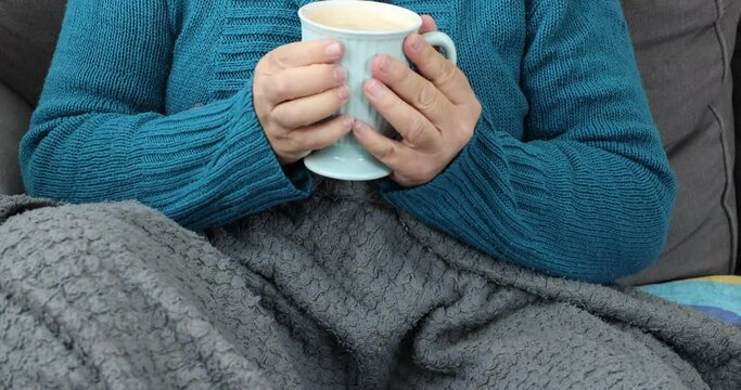 Female hands holding cup of coffee to warm up, very cold woman covered with gray blanket, wearing blue sweater, winter day, gas shortage and high prices. Concept of energy crisis in the European Union