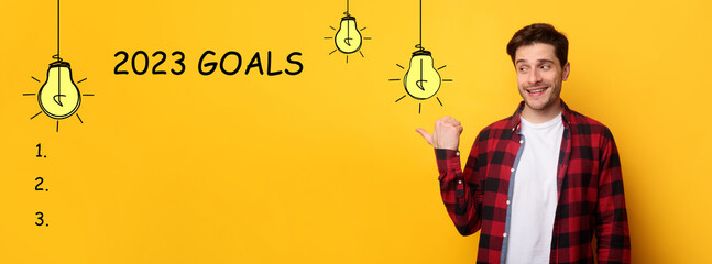 Cheerful millennial european guy point finger at goals 2023 with light bulbs, checklist with blank...