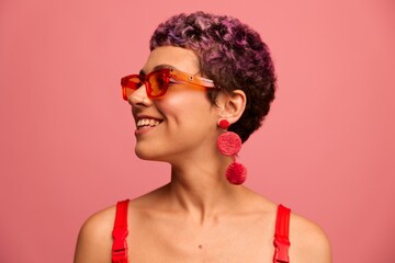 Fashion portrait of a woman with a short haircut in colored sunglasses with unusual accessories with earrings smiling on a pink bright background