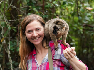 Young woman in shirt posing with small lemur on her shoulder, smiling, blurred jungle trees background © Lubo Ivanko