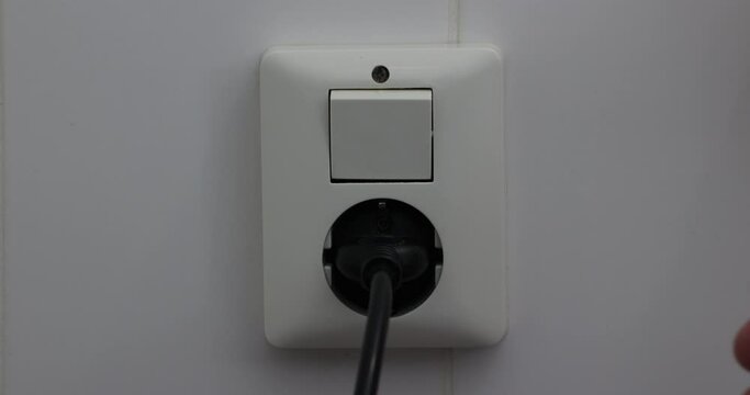 Hands of an adult woman unplugging a black cable from an outlet, a white tiled wall in a bathroom. Concept of saving electricity in the European Union energy crisis