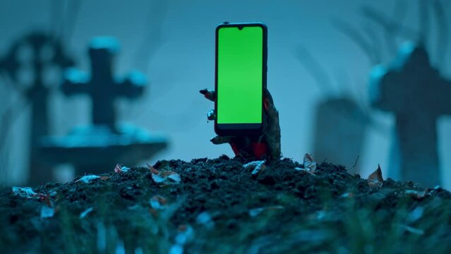 Zombie hand holds smartphone with green screen out of grave. Holiday event halloween concept.