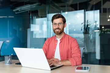 Portrait of mature businessman freelancer startup, bearded man smiling and looking at camera, business owner working inside modern office building wearing red shirt and glasses.