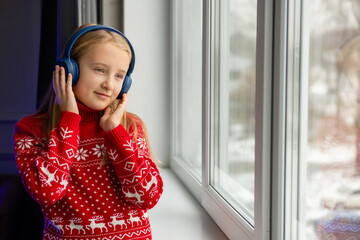 Little girl in red sweater listens to music or an audiobook in wireless headphones at the window of...