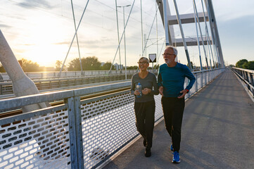 Cheerful active senior couple jogging together outdoors on the bridge. Healthy activities for elderly people.