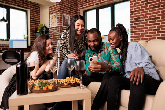 Multiethnic people group looking funny videos on smartphone screen, smiling, laughing, drinking wine, eating appetizers, cheese, bread sticks at apartment living room birthday party.
