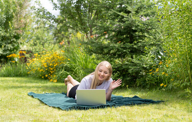 A young girl lying on the grass in the backyard with a laptop and communicates through a video call. 