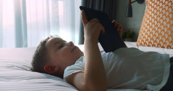Cute smiling child, kid boy using digital tablet technology device lying on bedroom alone. Small child hold pad computer surfing internet play game at home