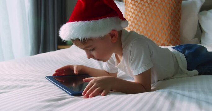 Small child hold pad computer surfing internet play game at home.Happy cute smiling child in Christmas hat Santa Clausa, kid boy using digital tablet technology device lying on bedroom alone.