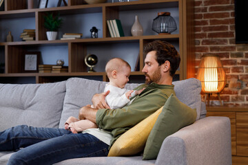 Dad plays with his little baby, on the couch in a cozy apartment. Fatherly love. Father hugs and kisses his little daughter.