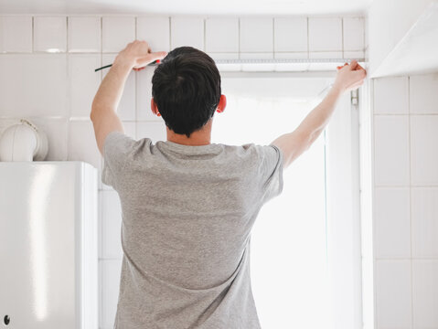 A young caucasian guy measures the width of a small window in the bathroom with a tape