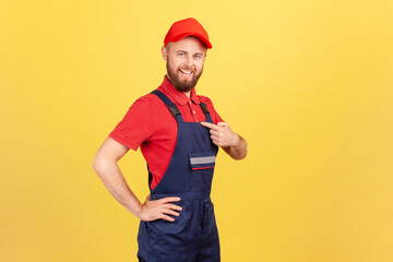 Side view of proud successful worker man standing and pointing at himself, bragging about the result of his work, wearing overalls and red cap. Indoor studio shot isolated on yellow background.