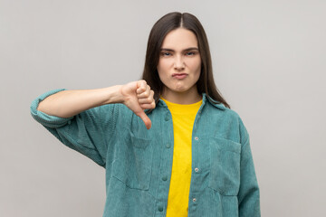Dissatisfied woman showing thumb down, disapproval sign, gesturing dislike to bad service, disagree with suggestion, wearing casual style jacket. Indoor studio shot isolated on gray background.