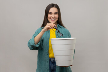 Smiling woman with dark hair standing holding trash bin and throwing out eyeglasses, improving eyesight after treatment, wearing casual style jacket. Indoor studio shot isolated on gray background. - obrazy, fototapety, plakaty