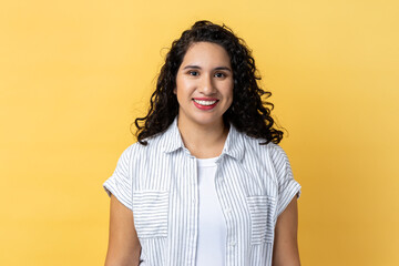 Portrait of cheerful positive woman with dark wavy hair looking at camera with toothy smile, expressing happiness, being in good mood. Indoor studio shot isolated on yellow background. - obrazy, fototapety, plakaty