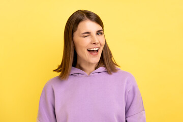 Obraz premium Portrait of positive optimistic woman standing and winking playfully, positive expression, looking at camera, wearing purple hoodie. Indoor studio shot isolated on yellow background.