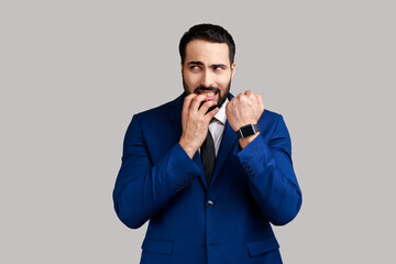 Nervous bearded man biting finger nails and looking at camera, showing his wrist clock, worried about deadline, wearing official style suit. Indoor studio shot isolated on gray background.