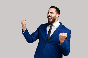Bearded man showing yes gesture and screaming celebrating his victory, success, dreams comes true, euphoria, wearing official style suit. Indoor studio shot isolated on gray background.